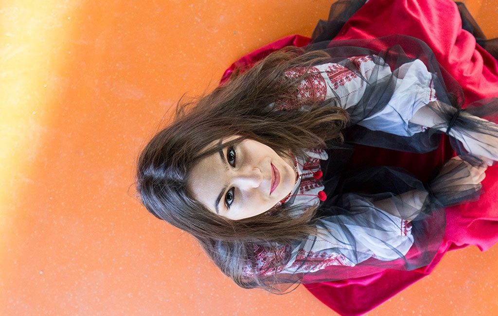 Model feminin asezat pe pozea, fotografiat de sus avand privirea in apaat si purtand un costum popular