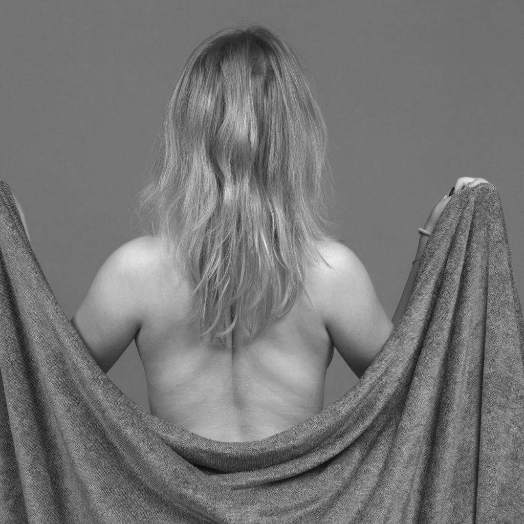 Sedinta foto boudoire cu o femeie cu spatele tinand o patura la nivelul spatelui ce ii acopera formele