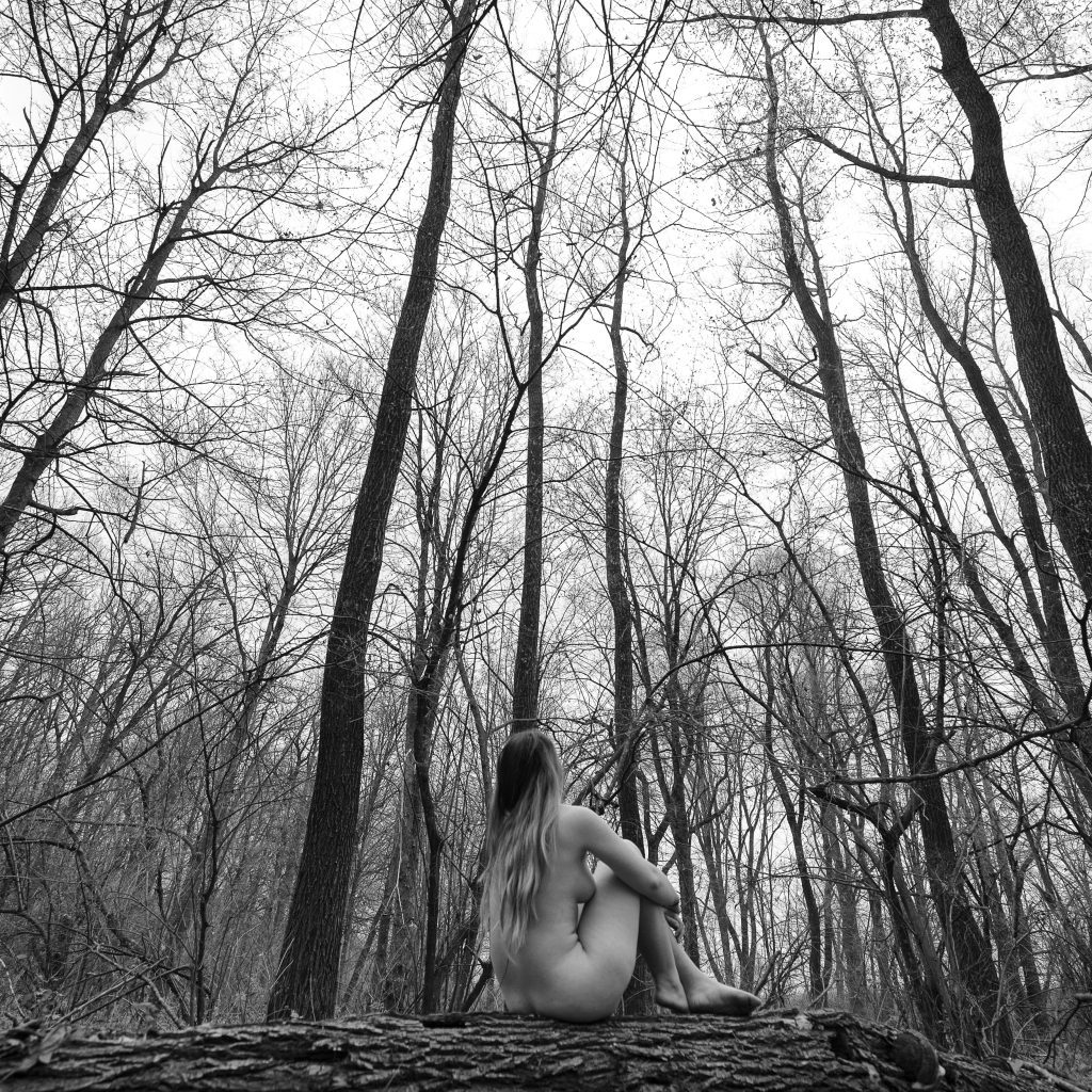 Fotografie conceptuala alb-negru: Model nud cu spatele in padure cu copaci goi, simbolizand forta naturii si a formei umane