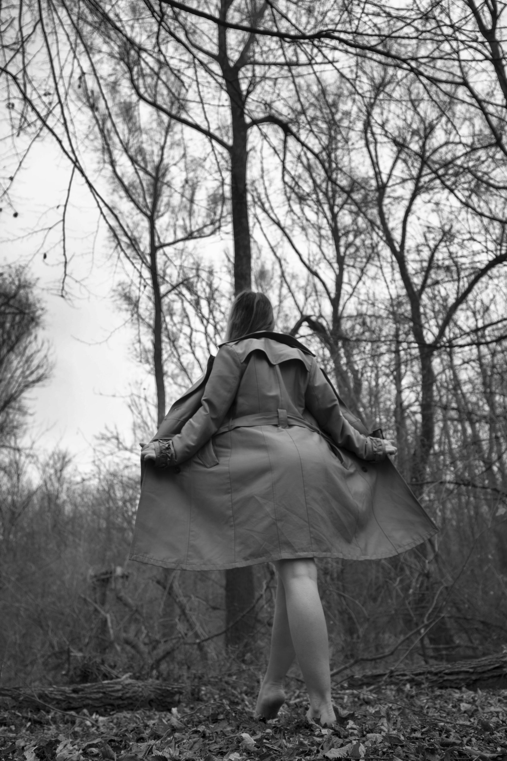 Fotografie fine art nude, model cu forme (curvy) în pădure. Toamnă, copaci goi, alb-negru, estetică cinematică cu trenci.