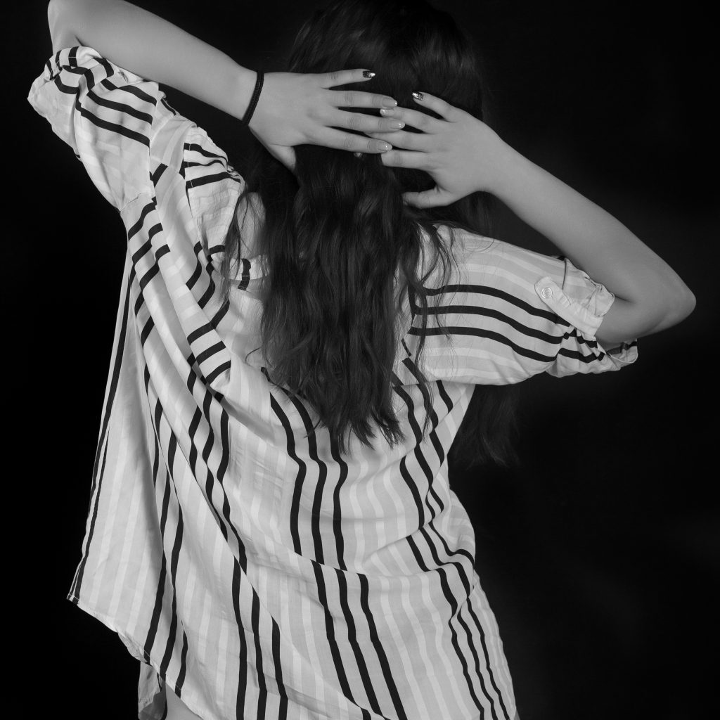 Fotografie boudoir artistică, alb-negru. Model feminin cu cămașă în dungi largă și lenjerie, brațe ridicate la ceafă, pe fundal de studio negru.