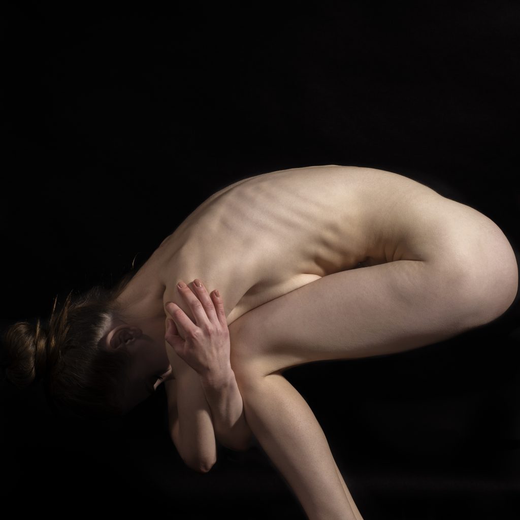 Fotografie fine art nud: siluetă feminină în postură yoga/dramatică pe fundal negru, contrast puternic lumină-umbră.