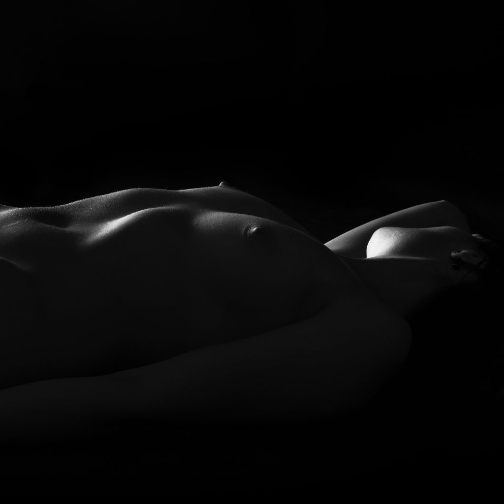 Portret boudoir cu contrast puternic: o femeie într-o poziție relaxată, corpul fiind modelat de jocul dramatic dintre umbre și lumină în studio.