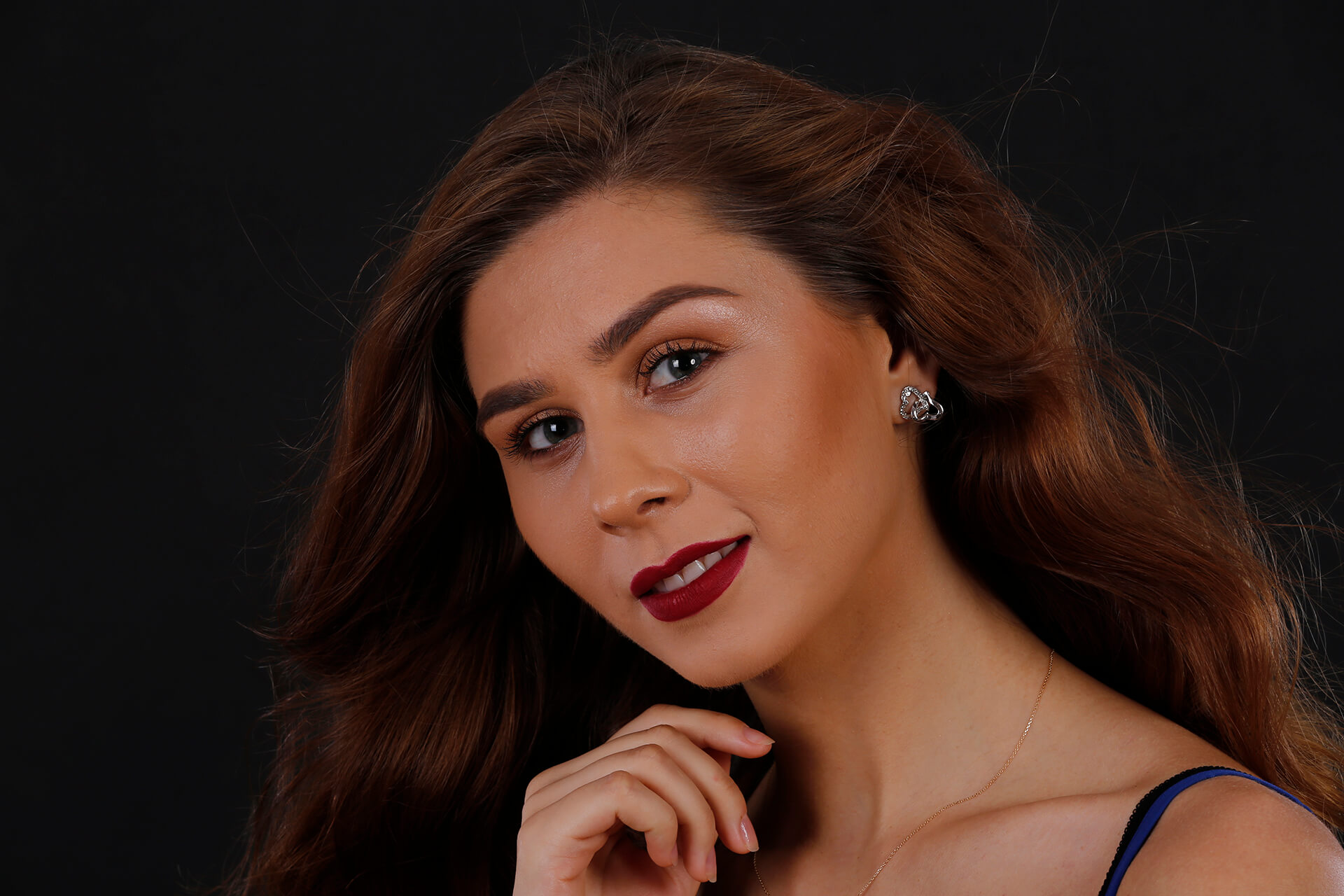 Portret feminin elegant realizat în studio foto din Iași. Femeie cu păr castaniu ondulat, ruj bordo și cercei discreți, pozând cu mâna la bărbie pe un fundal negru. Fotografie de branding personal de Andrian Photography.