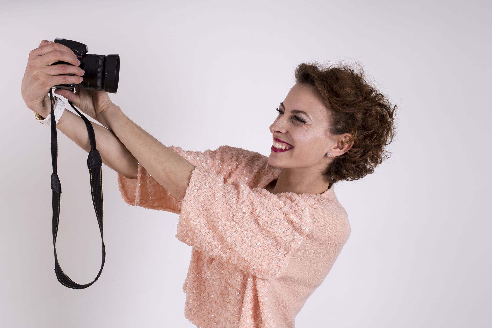 Portret dinamic realizat de Studio Foto Andrian în Iași, în care o tânără zâmbitoare ține un aparat foto DSLR ca și cum și-ar face un selfie, pe un fundal alb curat, sugerând o atmosferă relaxată la shooting.